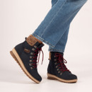 Person wearing blue jeans and dark navy winter boots with red lace-up fronts, metal eyelets, tan knit ankle cuffs, and beige rubber outsoles with rugged tread.