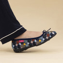 Side view of a person's foot wearing a cosy ballet-style slipper with a multi-coloured knit pattern and small round pom-pom details, finished with a navy blue trim and bow, and a pink "Lunar" brand tag on the side.