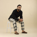 A man seated on a white wooden step stool against a neutral beige background. He is wearing a black long-sleeve top, cream and black check pyjama trousers with elasticated cuffs, and brown mule-style slippers with a distressed finish. His hands are clasped, and he looks relaxed and comfortable.