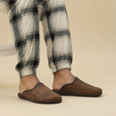 A person wearing brown mule-style slippers with a distressed suede-effect finish, dark brown sole, and visible front seam stitching. The slippers feature subtle embossed “dogz” branding on the side. The individual is dressed in cream and black checked pyjama bottoms with elasticated cuffs, standing on a neutral background.