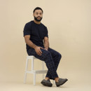 A man seated on a white step stool against a plain beige background. He is wearing a navy blue short-sleeve T-shirt, navy tartan pyjama trousers with light blue lines, and grey herringbone slippers with a check pattern and dark soles. His posture is relaxed, with hands resting on his knees.