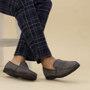 A person seated and crossing their legs, wearing grey herringbone slippers with subtle red and navy overcheck lines and dark grey trim. The slippers have black rubber soles and are styled for indoor comfort. The individual is also wearing navy blue pyjama trousers with a light blue and white windowpane check pattern.