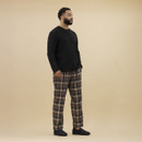 Man standing against a neutral background wearing a black long-sleeve top, beige and navy tartan check pyjama trousers, and navy blue check-patterned slippers. His hands are in his pockets and he is facing slightly to the right.