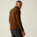 A man in a brown fleece jacket and black shorts looks over his shoulder. Neutral background, with a contemplative expression. Casual and relaxed tone.