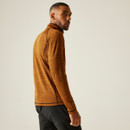 A man wearing a rust-coloured fleece and black trousers stands confidently, showcasing a casual yet stylish outfit.