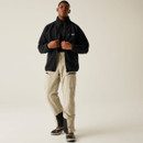 Front full-body shot of a man wearing the black fleece jacket unzipped over a base layer, revealing functional cargo trousers in beige with black boot overlays.