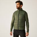 Male model wearing a Regatta Olive Night green padded bodywarmer with full front zip and high collar, styled over a green long-sleeve top and black hiking trousers.