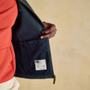 Close-up view of a man’s hand holding open the inside of a navy fleece gilet, revealing a large inner pocket and a stitched label reading “Joules – Built to last from town to country and shore to shore.” The gilet is worn over a red sweatshirt and cream trousers.