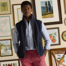 A man stands indoors in front of a wall decorated with an eclectic mix of framed artwork. He is wearing a navy blue sleeveless zip-up gilet over a light blue checked button-down shirt, paired with raspberry pink chinos. His hands are in his pockets and he has a calm, confident expression.