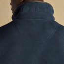 Close-up of the back of a navy fleece gilet featuring tonal embroidered "JOULES" branding across the top of the high collar. The garment includes brown trim detailing along the collar edge.