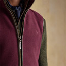 Close-up view of a burgundy-coloured fleece gilet with a brown zip and trim, worn over a dark green jumper and patterned navy neckerchief; features an embroidered hare logo and “JOULES” branded zipper.