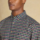 Close-up of a man wearing a green, navy, white, and red checked shirt with a button-down collar and chest embroidery. The shirt has a classic cut with wooden-look buttons and is styled with the top button undone.