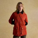 A woman wearing a red field jacket with black corduroy collar and cuffs. The jacket features gold popper buttons down the front, slanted chest pockets, and large flap pockets at the hips with gold studs. She stands with her hands in the pockets against a warm, textured beige background.