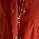 Detailed close-up of a women’s red field jacket, focusing on the central fastening. The gold-toned zip is engraved with the word 'JOULES' and accompanied by a matching pull tab. Surrounding the zip are two polished gold snap buttons and neatly stitched seams.