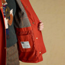 Close-up of a woman holding open a red field jacket to reveal its interior. The inside features a classic check lining in navy, cream, and rust tones. A stitched fabric label with the Union Jack and Joules branding is visible on the inner pocket, with text highlighting British design and quality. The woman wears a forest green jumper with an embroidered fox motif and brown check trousers.