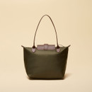 Rear view of a women’s olive green tote bag with brown faux leather accents. The bag features two slim shoulder straps and a back view of the flap closure. It is shown standing against a soft beige background.