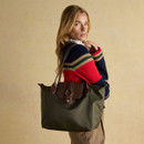 A woman with long blonde hair carries an olive green tote bag with brown faux leather detailing over her shoulder. She wears a bold red, navy, and green striped jumper with checked trousers, standing against a neutral beige backdrop.