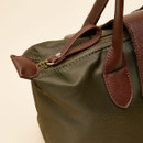 Close-up of the top corner of a women’s olive green tote bag, showing the brown faux leather handle attachment, matching zip pull tab with a metal ring and rivet detail, and the fabric’s fine stitching.