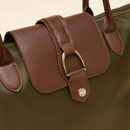 Close-up of a women’s tote bag showing the brown textured faux leather flap with a central strap looped through a gold-tone D-ring and secured with a gold popper embossed with the 'Joules' brand name. The body of the bag is olive green with matching brown handles.