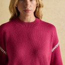 Close-up of a woman wearing a bright pink knitted jumper with a ribbed crew neckline and white zigzag stitching detail across the upper sleeves. Her blonde hair is loose, and she wears small gold hoop earrings. The background is a soft beige tone.