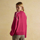 Rear view of a woman wearing a bright pink jumper with white zigzag stitching along the shoulder seams. The jumper has a ribbed hem and cuffs, and is paired with checked trousers. She is standing against a soft beige backdrop, looking back over her shoulder.