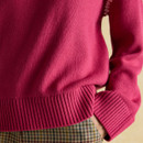 Close-up of the hem and cuff detail on a bright pink knitted jumper, worn over high-waisted checked trousers. The jumper features ribbed edging at the bottom and sleeves, with the model's hand tucked casually into the pocket.