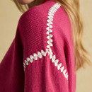 Close-up detail of a bright pink knitted jumper showing white zigzag embroidery running across the shoulder and down the sleeve. The texture of the knit and stitching is clearly visible, with part of the model’s blonde hair in the background.