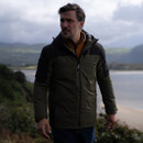 A man wearing a dark green jacket walks along a coastal path. The background shows a cloudy sky, mountains, and a glimpse of the sea, creating a serene, contemplative mood.