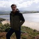 A person stands on a grassy cliff overlooking a serene beach and calm water under a cloudy sky. They wear a hooded jacket, conveying a relaxed, contemplative mood.