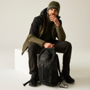 Man wearing outdoor gear, including a beanie and jacket, sits pensively on a white cube. He rests one hand on a black hiking backpack.