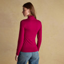 Back view of a woman wearing a berry-coloured long-sleeve top with a fitted silhouette and ruffled high neckline, paired with mid-wash blue jeans. She stands against a beige textured background, looking over her shoulder.