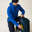 A person in a blue jacket and beanie kneels while placing a green water bottle into a blue backpack. The setting is neutral, suggesting a focus on outdoor gear.