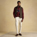 Full-body image of a man wearing a burgundy jumper with an embroidered car design, layered over a dark green and navy shirt, and paired with cream-coloured trousers, brown loafers, and an olive green zip-up gilet. He stands with one hand in his pocket against a beige backdrop.