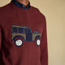 Close-up of a burgundy knitted jumper featuring an embroidered 4x4 vehicle motif in navy, mustard, and cream tones, worn over a dark striped shirt with buttoned collar.