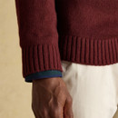 Close-up of a man’s lower arm and hand wearing a burgundy knitted jumper with ribbed cuffs and hem, layered over a dark green and navy shirt, paired with cream-coloured trousers.