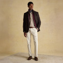 Man dressed in a smart-casual outfit featuring a dark brown checked blazer with a burgundy lining, layered over a beige check shirt and cream trousers. He accessorises with a patterned neck scarf and brown tasselled loafers, standing on a neutral-toned studio backdrop.