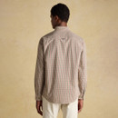 Back view of a man wearing a traditional checked shirt in cream, green, and red tones, paired with off-white trousers. The shirt features a classic fit with a curved hem and pleated back yoke detail, set against a neutral background.