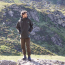 Man standing on rocky terrain wearing a forest green Monsoon Classic waterproof jacket with hood, shown in an outdoor mountain setting.