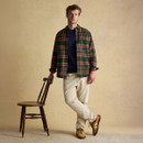 Male model standing in a relaxed pose, lightly holding the backrest of a wooden spindle chair with one hand. He is wearing a dark green, mustard, and claret tartan shirt over a navy top, paired with cream corduroy trousers and tan trainers. The backdrop is plain and beige, creating a warm, rustic studio setting.