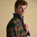 Close-up three-quarter profile of a male model wearing a green, mustard, and claret tartan shirt with a denim-look internal collar lining. The model holds the shirt partially unbuttoned at the neck and looks slightly over his shoulder against a plain beige backdrop.