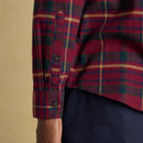 Close-up of the cuff and lower sleeve of a red tartan shirt with green, navy, and mustard checks; features buttoned cuff and placket, with part of a dark navy trouser leg visible below.
