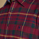 Close-up of a red flannel shirt with a tartan check pattern in green, yellow, and navy. The shirt features a button-up front, a front pocket with an embroidered navy hare logo, and a contrast chambray lining on the collar.