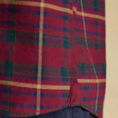 Close-up of the side seam and hem of a red tartan shirt with green, navy, and mustard checks; features a reinforced gusset detail at the curved hem, worn over dark navy trousers.