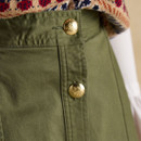 Close-up of the waistband and front fastening of an olive green skirt, featuring shiny gold metal buttons embossed with a crest motif. The textured knit of a Fair Isle-style vest in warm red, navy, and beige tones is partially visible above.