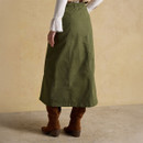 Rear view of a woman wearing a long olive green A-line skirt with visible seam detailing and an elasticated waistband at the back. She is dressed in a white blouse with flared cuffs and brown suede knee-high boots, standing against a neutral beige background.