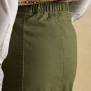 Close-up of the back waistband of an olive green skirt, showing a gathered elastic panel for added comfort and fit. The wearer is dressed in a white blouse with gold cuff buttons and a patterned knit vest in muted tones.