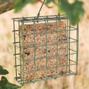 A square suet block filled with seeds is enclosed in a green metal feeder, hanging outdoors from a tree branch, with blurred greenery in the background.