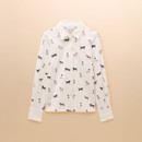 Flat lay of a long-sleeved white shirt with a classic collar and button-down front, featuring an all-over print of various dog breeds in shades of brown and black. Displayed against a soft beige background.