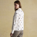 Rear view of a woman wearing a white shirt adorned with a playful dog print in neutral tones. The shirt has a structured collar and buttoned cuffs, paired with high-waisted brown and olive check trousers. She stands in front of a soft beige background, turning slightly to the side.