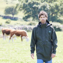 Outdoor lifestyle shot of a man wearing a forest green waterproof smock jacket in a grassy field with cows grazing in the background.